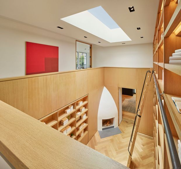 A bright, carefully detailed interior view across a split-level library or reading space. Warm timber wall panelling and extensive built-in open shelving line the right side and lower walls, filled with books and objects. A timber balustrade overlooks a lower level with a sculptural white arched fireplace and herringbone timber flooring. A large skylight, recessed ceiling lights, a red feature artwork, and a leaning library ladder complete the contemporary space.