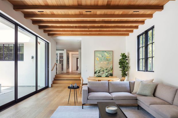 A bright, open-plan living room with a large L-shaped sectional sofa, timber flooring, and a dramatic exposed timber-beam ceiling. Full-height black-framed glazed sliding doors open to an adjoining outdoor space, while tall black-framed windows bring in natural light. A dining table, artwork, indoor plant, and a hallway or stair area are visible in the background. The room has a warm, contemporary character with strong natural timber detailing.