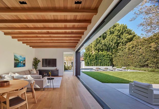 Open-plan living and dining area with a warm timber-lined ceiling and exposed timber beams, pale timber flooring, and large retractable floor-to-ceiling glazing opening directly to a lawn and poolside terrace. The space includes a neutral upholstered sofa, timber dining table and chairs, and understated contemporary furnishings. A strong indoor-outdoor connection dominates the composition.