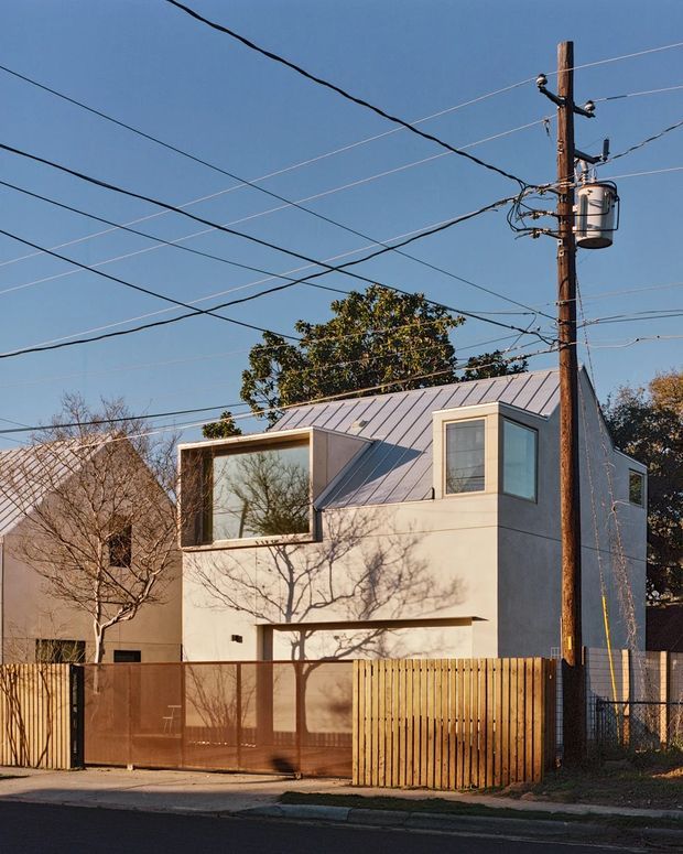 Street-facing view of a compact contemporary urban residence with a pale rendered facade, standing-seam metal pitched roof, projecting upper-level window, and large glazed opening. A timber slat fence and translucent metal driveway gate enclose the property, while overhead utility wires and a timber power pole dominate the foreground.