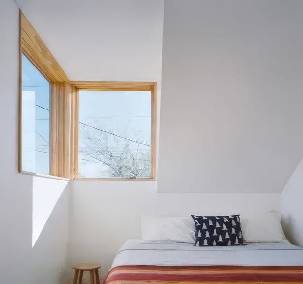 Bright, minimalist bedroom beneath a sloping ceiling, featuring a low bed with layered white and rust-toned bedding, a small wooden stool, and a distinctive corner window with exposed light timber framing. The room has white walls, abundant daylight, and a calm, pared-back aesthetic.