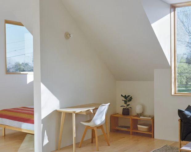 Bright, compact bedroom or studio sleeping area beneath a sloping ceiling, with a single bed, small timber desk, white molded chair, mid-century timber sideboard, indoor plant, and large timber-framed windows. The room has white painted walls, pale timber flooring, and strong natural daylight.