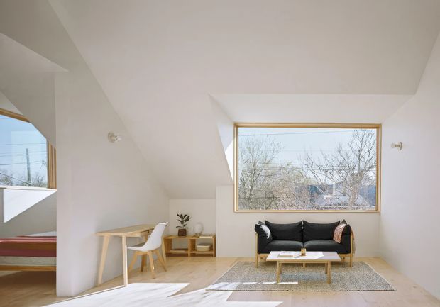 Bright minimalist living room with white sculptural walls and ceiling planes, pale timber flooring, and a large timber-framed picture window. A compact black sofa, low coffee table, neutral rug, and simple light-wood desk with white chair furnish the space. An adjacent sleeping area is partially visible through an opening on the left.