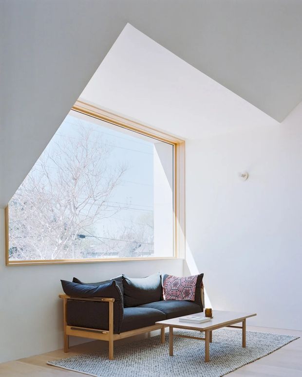 Bright minimalist living room with a black upholstered mid-century-style sofa in a timber frame, a low timber coffee table, and a textured area rug. A large timber-framed picture window brings in abundant natural light, while the sharply angled ceiling and wall create a distinctive geometric architectural feature. The palette is predominantly white with light timber and dark upholstery accents.