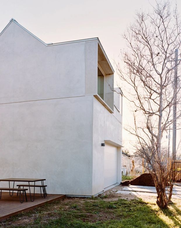 Exterior view of a minimalist two-storey white rendered house with a simple gabled roof form, recessed upper-level balcony or window opening, and a large garage door. A timber deck with an outdoor dining table sits beside the house, with a sparse winter garden and fencing visible around it.