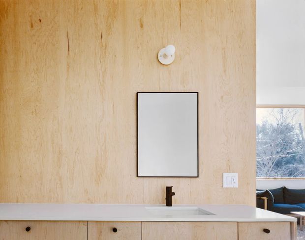 A minimalist bathroom vanity with a full-height pale timber wall panel, white benchtop and integrated basin, dark bronze or black tapware, frameless rectangular mirror, and simple wall light. A window and part of an adjacent seating area are visible at right.