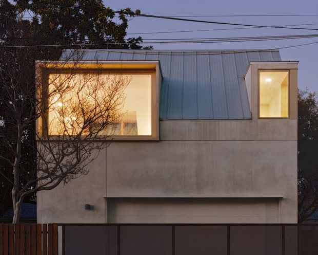 Twilight street-level view of a compact contemporary multi-storey house facade. The building has a smooth pale rendered lower facade, a prominent standing-seam metal pitched roof, two projecting illuminated upper-level window bays, and an integrated garage concealed behind a dark front gate. Overhead utility wires and mature trees frame the restrained urban residential exterior.