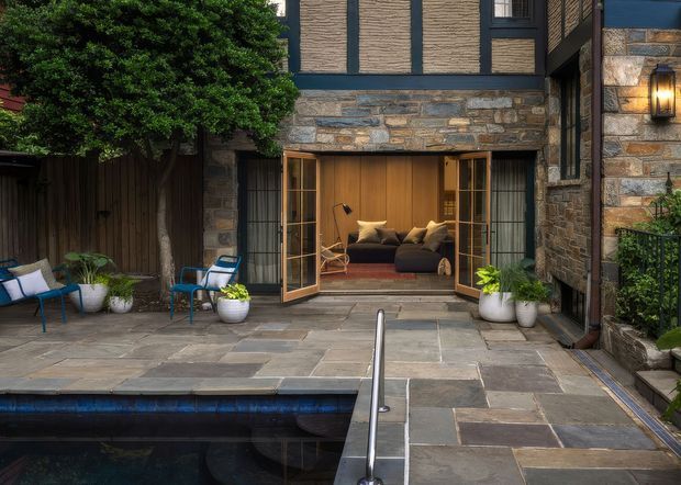 A sheltered stone-paved courtyard and pool terrace viewed toward a contemporary stone-clad building. Open timber-framed glazed doors reveal a warmly furnished living room, while dark timber or painted detailing, natural stone walls, outdoor seating, potted plants, and a narrow swimming pool create a refined indoor-outdoor setting.