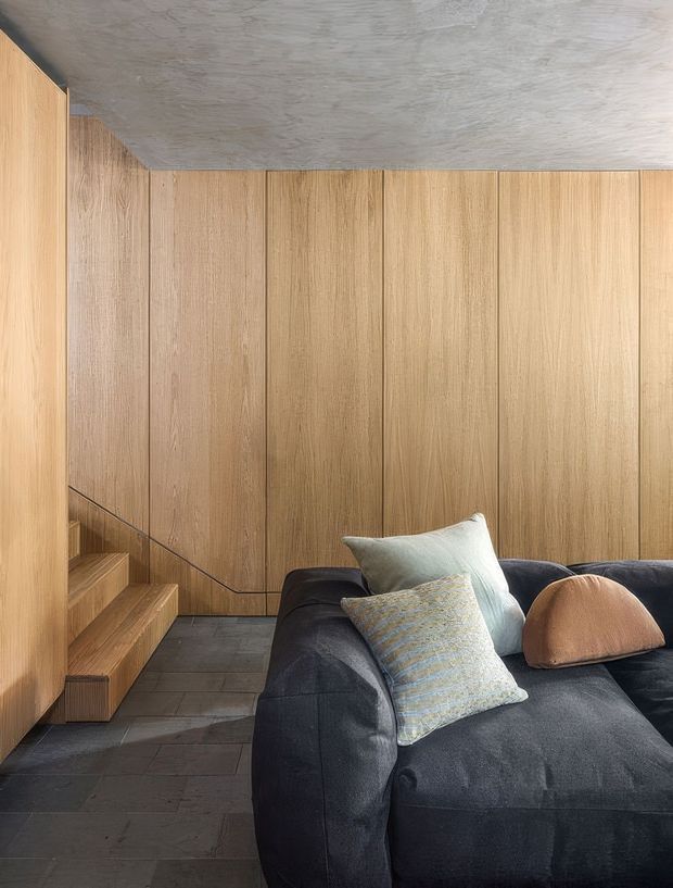 A compact, refined living area featuring extensive vertical timber wall panelling and integrated timber stair cabinetry. A dark charcoal sectional sofa with layered cushions sits on a dark slate-like tiled floor, beneath a textured grey concrete ceiling. The space has a warm, tactile, minimalist character with strong built-in joinery and concealed stair detailing.