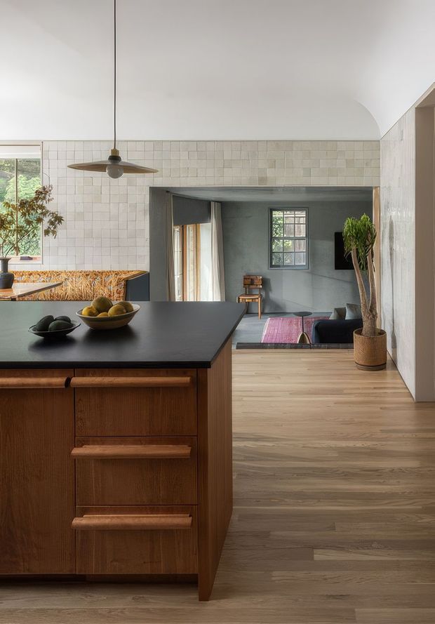 A warm, refined open-plan kitchen looking toward a connected living area. The kitchen features a dark matte benchtop, rich timber island cabinetry with sculpted drawer pulls, a tiled backsplash, a suspended ceiling fan/light, and timber flooring extending through the living space. A tiled opening frames the darker lounge, which contains seating, a television, and a small table. Natural light enters through large windows, while potted plants and fruit bowls add organic accents.