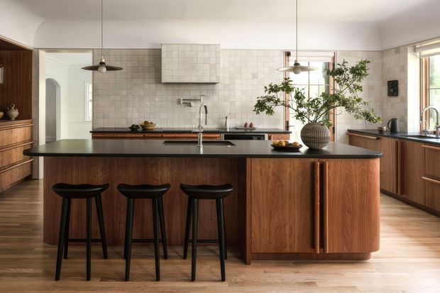 A warm, refined kitchen centred on a large dark stone island with seating for three. The island and surrounding cabinetry feature rich mid-to-dark timber joinery with distinctive vertical pulls, complemented by a white square-tiled splashback, integrated rangehood, timber flooring, and brushed metal fixtures. Pendant lights, open shelving, and a large indoor tree add character to the contemporary space.