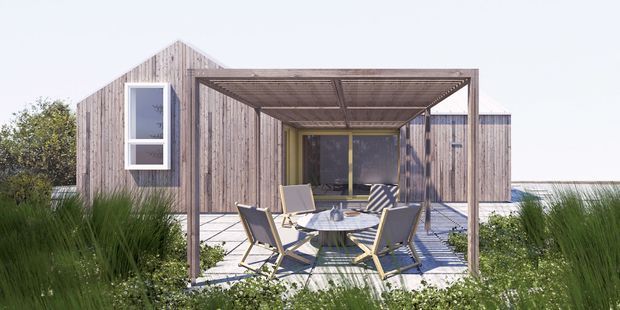 Exterior view of a compact modern, single-storey house with weathered timber cladding and a pitched gable roof. A flat-roofed timber-framed pergola shelters an outdoor dining and lounge area on a paved terrace, surrounded by ornamental grasses and low planting. Large glazed doors connect the terrace to the interior.