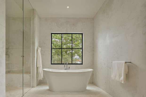 A serene, minimalist bathroom featuring a freestanding white bathtub beneath a black-framed grid window, a glass-enclosed shower, pale marble-look wall and floor surfaces, and simple white towel rails. Natural greenery is visible outside the window.