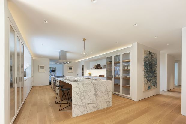 Bright open-plan kitchen with a large marble-look island, integrated seating, white cabinetry, glass-front display storage, timber flooring, and recessed ceiling lighting. Full-height glazed doors or windows line the left side, while a hallway and artwork are visible to the right. The space has a refined, contemporary feel with warm timber and white finishes.