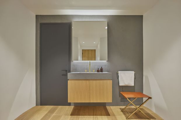 Compact contemporary bathroom with a wall-mounted light-wood vanity, grey stone-look benchtop, large rectangular mirror, dark grey door, recessed ceiling lights, towel rail, and a small folding stool. Light timber flooring and neutral walls create a clean, restrained interior.