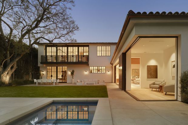 Twilight exterior view of a refined courtyard residence with a flat-roofed contemporary wing opening through expansive glazed doors to a warmly lit living area. The property includes a manicured lawn, swimming pool, paved terrace, dark-framed windows, a balcony, and a tiled low-pitched roof on the adjoining two-storey section. The architecture combines clean modern volumes with restrained traditional Mediterranean-influenced roof detailing.