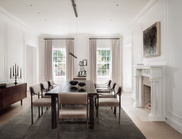 A refined formal dining room with a long dark timber dining table surrounded by upholstered dining chairs. The bright white room features tall multi-pane windows with full-length striped curtains, detailed wall panelling, ornate fireplace mouldings, a large neutral rug, a sculptural pendant light, and a dark timber sideboard with candlesticks. The overall composition is elegant, balanced, and traditional with contemporary furnishings.