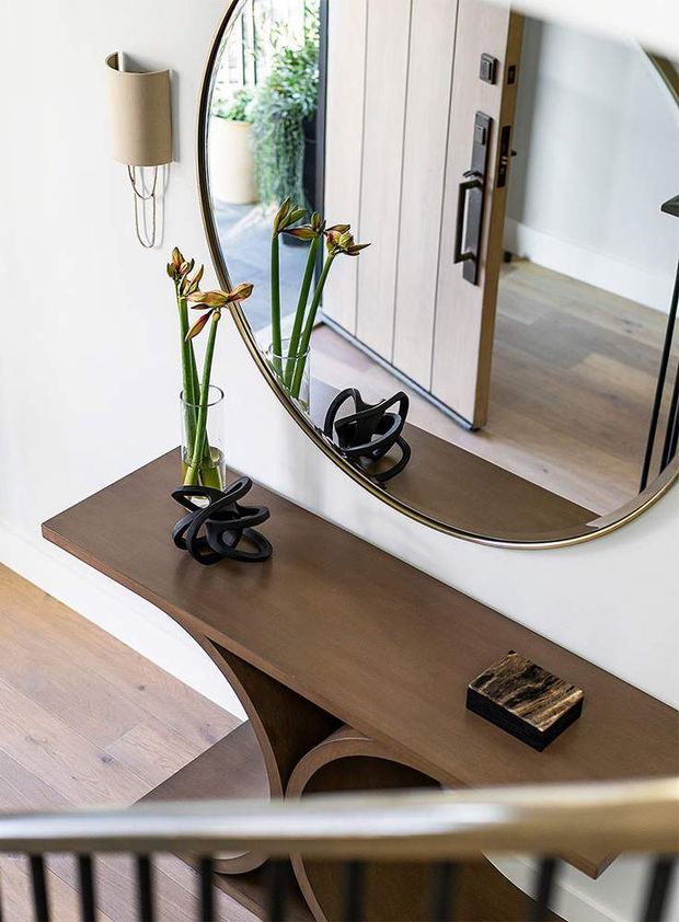 A refined residential entryway with a dark timber console table beneath a large round brass-edged mirror. Sculptural black objects, a glass vase with flowers, and a wall-mounted cylindrical light decorate the console. The mirror reflects a timber-framed doorway, pale walls, timber flooring, and greenery outside. A dark metal stair railing crosses the foreground.