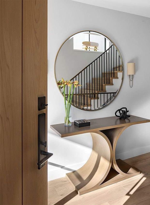 A refined residential entryway viewed through an open timber door. The space features a sculptural console table with a dark stone-like top and curved light-wood base, a large round mirror, a slim wall sconce, timber flooring, and a black metal stair balustrade reflected in the mirror. Natural sunlight creates strong shadows across the floor and console.