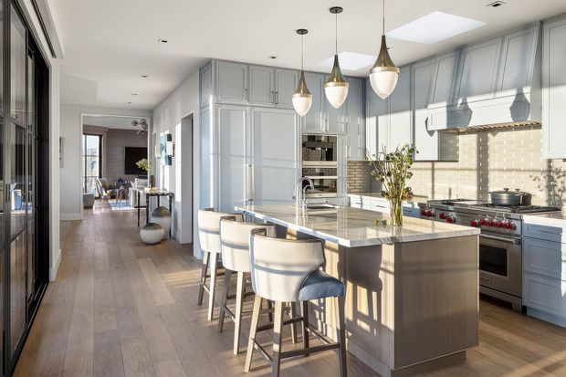 Bright, spacious contemporary kitchen with a large central island, integrated seating for four, pale blue-grey shaker-style cabinetry, a light stone benchtop, and a beige brick-tile splashback. Pendant lights hang above the island, while a substantial stainless-steel range and hood form the cooking zone. Timber flooring runs through the kitchen and adjoining hallway, with strong natural light entering from glazing and a skylight.