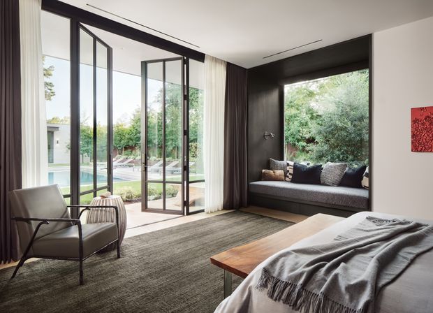 A refined contemporary bedroom with floor-to-ceiling black-framed glazed doors opening toward a landscaped pool area. The room features a built-in dark window seat with cushions, a low timber coffee table, a large textured rug, a modern armchair, layered curtains, and a partially visible bed with a grey throw. The palette is neutral and moody with strong indoor-outdoor connection and abundant natural light.