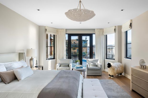 Bright, elegant bedroom with a partially visible upholstered bed, layered neutral bedding, a pair of armchairs, a small central table, a bench, and a light wood dresser. The room has large black-framed windows and a glazed balcony door with city views, pale curtains, light timber flooring, a sculptural pendant light, and restrained neutral styling.