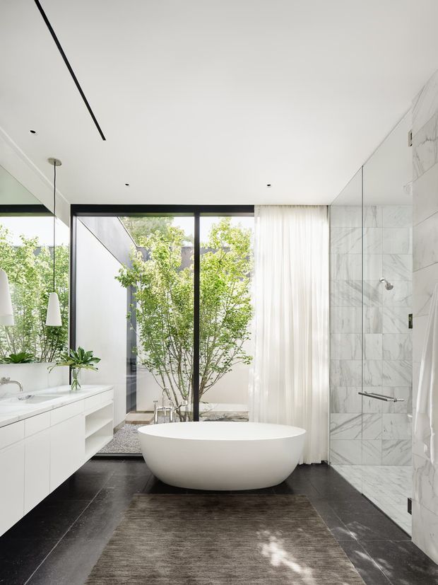Bright, refined contemporary bathroom with a freestanding white oval bathtub centered in the room. The space features a large marble-tiled shower wall, dark stone-look flooring, a white floating vanity with integrated basin, frameless mirror surfaces, sheer curtains, black-framed glazing, and a planted courtyard visible beyond the glass. Minimal pendant lights and clean-lined joinery create an airy, luxurious atmosphere.