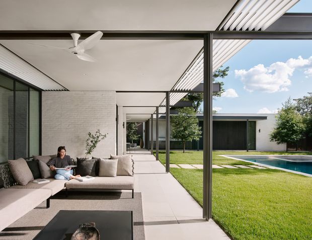 A modern single-storey residence with a covered outdoor living terrace overlooking a manicured lawn and swimming pool. The terrace features a low-profile sectional sofa, dark coffee table, slim metal posts, a flat roof with linear louvred screening, and extensive glazing. White painted brick walls, pale concrete paving, dark frames, and an adjacent pool create a clean indoor-outdoor composition.