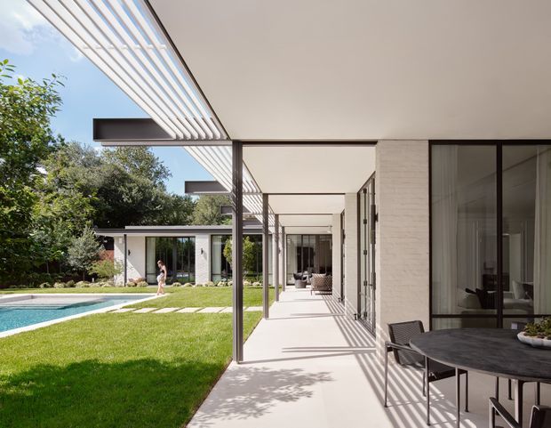 A bright modern single-storey home arranged around a landscaped courtyard and swimming pool. The covered outdoor passage features a long white roof canopy with slender dark metal posts and linear pergola-like slats, opening onto a lawn and pool. The facade combines white painted brick, extensive dark-framed glazing, and pale concrete or stone paving. Outdoor dining furniture occupies the foreground, while a separate pavilion-like wing is visible across the garden.