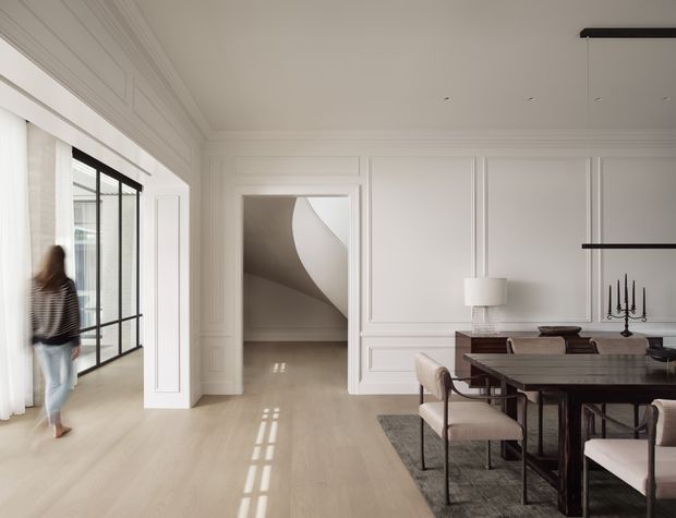 A refined, bright open-plan dining area with a dark rectangular dining table and upholstered chairs, framed by white decorative wall panelling and detailed ceiling mouldings. A large black-framed glazed opening brings in strong natural light, while a sculptural curved staircase is visible through the central doorway. Minimal black linear pendant lighting and restrained furnishings create a contemporary, elegant interior.