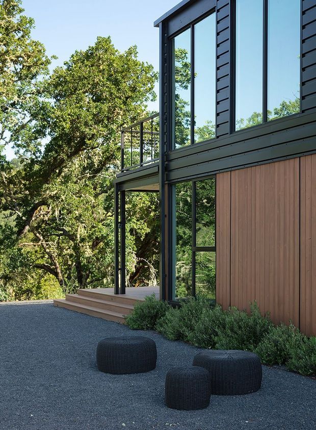 A modern multi-level house exterior set within mature trees, featuring a dark metal-clad upper facade, extensive black-framed glazing, warm vertical timber wall cladding, and a raised timber deck with steps. A dark gravel outdoor area with three black woven ottomans sits beside low planting.