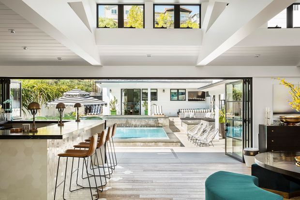 A bright, expansive indoor-outdoor entertaining space centred on a swimming pool courtyard. The interior features a large kitchen island with bar seating, extensive folding or sliding glass doors opening to the pool, white structural beams and ceiling panelling, and a mezzanine or upper-level clerestory with black-framed windows. Outdoor loungers and a pool are visible beyond, while a curved dark dining table and teal upholstered seating occupy the foreground.