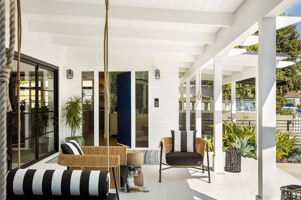Bright, airy covered outdoor lounge or sunroom with a white-painted timber structure, exposed beams, and broad open sides. The space includes a hanging rope swing with a black-and-white striped cushion, woven lounge seating, a mid-century-style armchair, potted plants, and a glimpse of the adjacent interior through black-framed glazed doors. The overall aesthetic is relaxed coastal with strong monochrome accents.