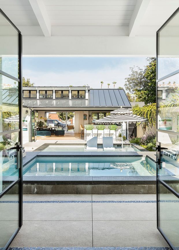 A bright architectural view from inside a contemporary home through fully opened black-framed glazed doors toward a private courtyard pool. The pool sits directly outside on a tiled terrace, with white loungers, a striped outdoor umbrella, bar seating, tropical planting, and a modern pavilion-like building with a standing-seam metal roof beyond. White exposed ceiling beams and extensive glazing frame the view.