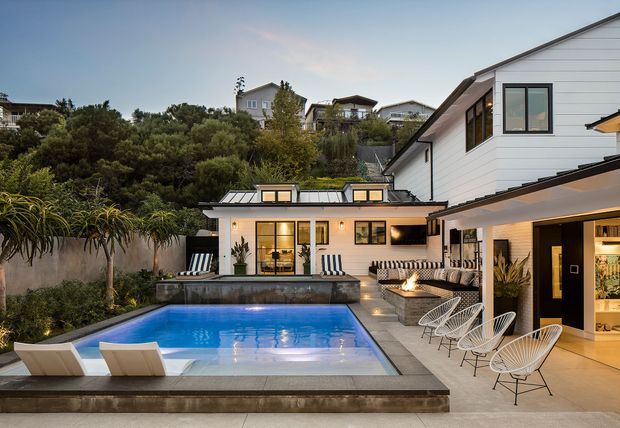 A dusk exterior view of a contemporary multi-level residence centered around a rectangular swimming pool and paved entertaining terrace. The home has white horizontal cladding, dark-framed windows, a low-pitched roof, extensive glazing, and an adjoining outdoor lounge with striped seating and sculptural wire chairs. Lush planted screening and neighboring hillside homes form the background.