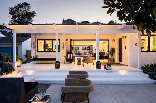 A dusk exterior view of a contemporary white-painted brick single-storey home opening onto a broad covered patio and outdoor entertaining area. The patio has a flat pergola-style roof, built-in lighting, outdoor seating, a fire feature, tiled steps, and a clear view into the warmly lit living and dining spaces. Layered landscaping and neighbouring homes surround the terrace.