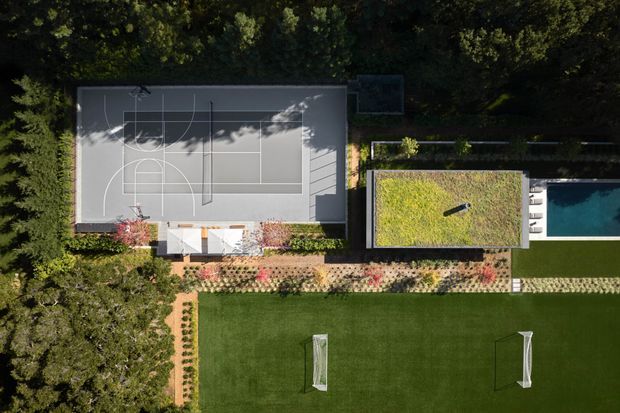 Aerial view of a landscaped residential recreation area featuring a full-size outdoor basketball court, a rectangular swimming pool beside a planted green-roof structure, and a large manicured lawn with two soccer goals. Dense trees and patterned garden planting surround the sports and leisure spaces.