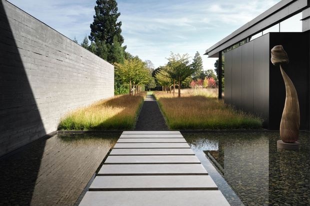 A minimalist contemporary courtyard landscape with a narrow stepping-stone path crossing a shallow reflecting pool. A tall raw concrete boundary wall lines the left side, while a dark-clad low pavilion with a projecting roof and large black opening sits to the right. Ornamental grasses, clipped planting, mature trees and a sculptural timber form create a serene, strongly geometric composition.