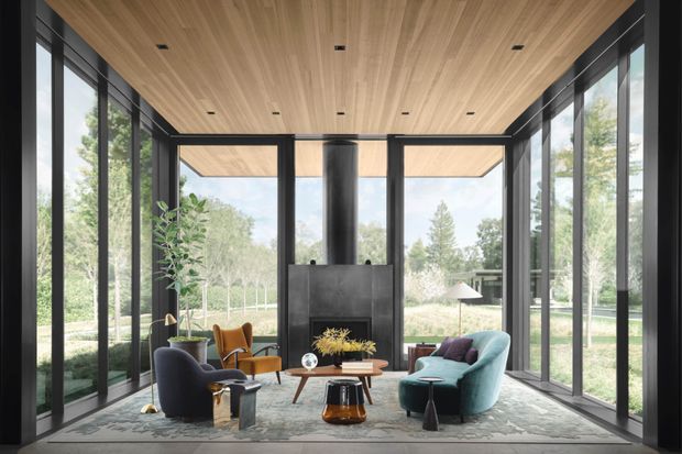 A refined, light-filled living room enclosed by floor-to-ceiling black-framed glazing on three sides. The room features a warm timber-lined ceiling, dark fireplace surround and chimney, a sculptural teal sofa, lounge chairs, a central coffee table, and a large patterned rug. Views of landscaped grounds are visible through the glazing.