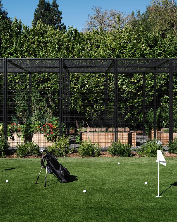 A landscaped outdoor golf practice area with manicured artificial turf, a putting hole and flag, golf balls, and a golf bag. Behind it is a tall black metal mesh enclosure containing raised garden beds and dense greenery, creating a screened garden or training enclosure.