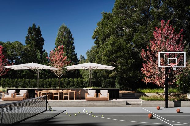 Outdoor residential entertainment area with a full-size basketball half-court in the foreground, adjacent to a landscaped patio with outdoor dining, lounge seating, umbrellas, and mature trees with pink blossoms. The space has a refined contemporary resort-like character and appears designed for recreation and entertaining.