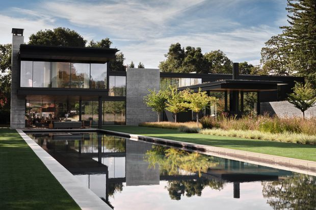 Exterior view of a sophisticated low-profile modern residence with dark metal framing, expansive floor-to-ceiling glazing, a textured grey masonry feature wall, and multiple flat-roofed volumes. A long reflecting-style swimming pool and manicured landscaped garden occupy the foreground, with mature trees surrounding the property.