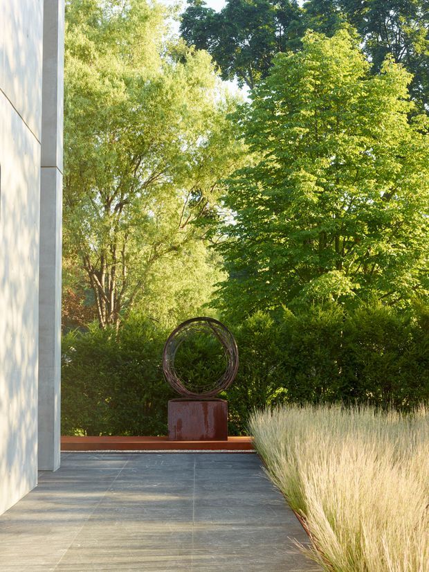 A contemporary garden courtyard or terrace viewed beside a pale rendered building wall. Large dark grey rectangular paving slabs lead toward a circular metal sculpture on a rust-toned plinth, bordered by clipped hedges and tall ornamental grasses, with mature leafy trees beyond. The scene is softly sunlit and strongly connected to the surrounding landscape.