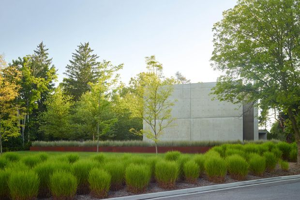 Exterior architectural photograph of a minimalist concrete building partially screened by mature trees and layered ornamental grasses. The simple rectangular volume has a smooth pale concrete facade with a dark vertical recessed section, set within a carefully landscaped garden beside a paved road or walkway.