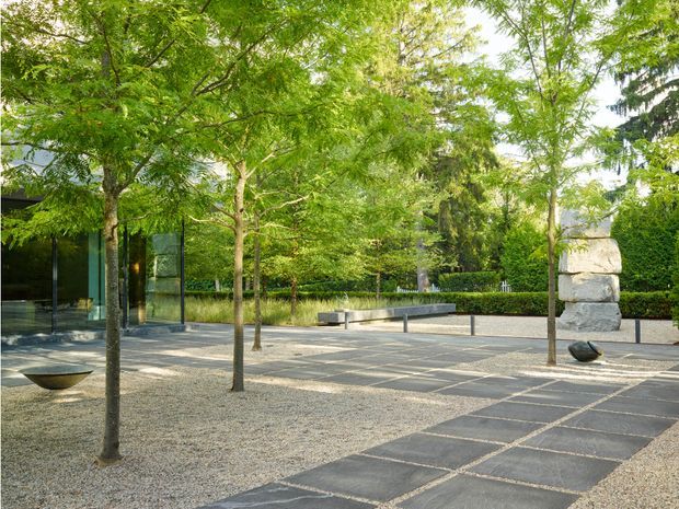 A landscaped architectural courtyard or garden with rows of leafy trees, pale gravel ground cover, dark rectangular stone paving, low minimalist concrete seating or retaining elements, clipped hedges, and a partially visible modern glazed building. A large stacked-stone wall feature anchors the right side of the scene. The design is calm, geometric, and contemporary.