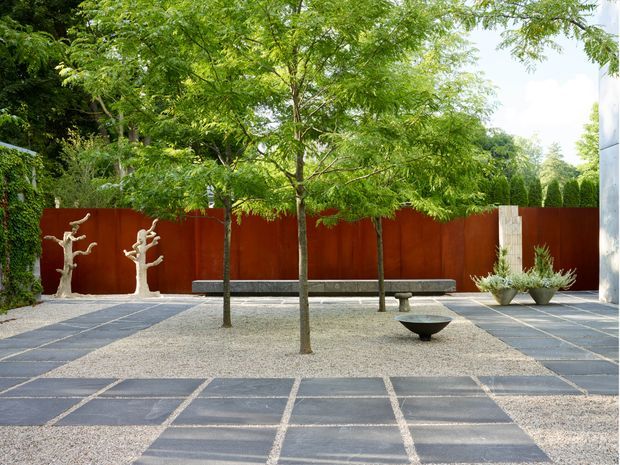 A landscaped courtyard or garden terrace with a geometric grid of large dark paving slabs set in pale gravel. Young leafy trees, a long dark bench, sculptural pale branch forms, planters, and a rust-red metal privacy screen create a calm, minimalist outdoor composition.