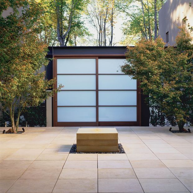 A minimalist courtyard or driveway with large pale square paving tiles leading to a dark-framed, frosted-glass garage or entrance door. Warm timber framing, clipped trees, dense planting, and a low rectangular stone feature set into a gravel recess create a calm, Japanese-influenced garden composition.