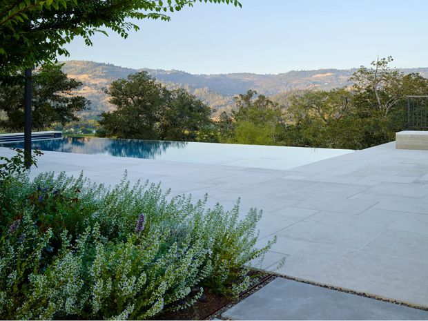 A landscaped outdoor terrace with a large pale stone-paved pool deck and an infinity-edge swimming pool overlooking wooded hills and a broad valley. Dense ornamental planting borders the foreground, while the pool and terrace create a calm, elevated outdoor setting.