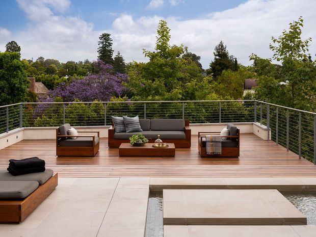 A spacious elevated outdoor terrace or rooftop deck overlooking lush mature greenery and neighbouring rooftops. The terrace has warm timber decking, pale stone-paved areas, a low linear water feature or reflecting pool, sleek metal cable railings, and a contemporary outdoor lounge arrangement with sofas, armchairs, cushions, and a coffee table. The scene is bright and relaxed with a refined contemporary resort-like atmosphere.