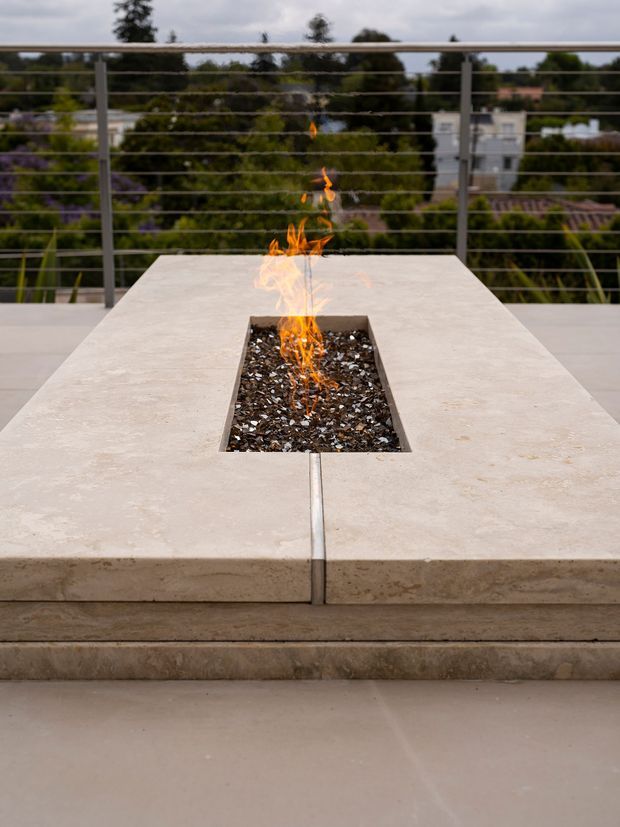 Close-up view of a rooftop or elevated outdoor terrace featuring a built-in rectangular fire feature set into a large pale stone or concrete table. The fire pit is filled with dark decorative stones and surrounded by broad slab surfaces, with a metal cable balustrade and leafy suburban surroundings beyond.